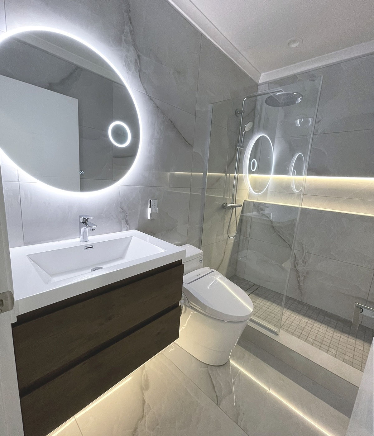 Modern bathroom renovation with LED backlit mirror, marble tile shower, and floating wood vanity by JSP Construction